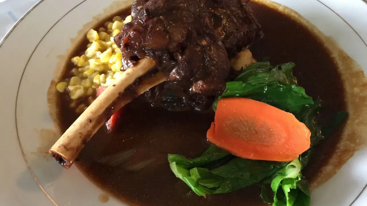 Delicioso lechazo guisado con hueso, acompañado de maíz tierno, zanahoria y verduras verdes en un plato blanco.