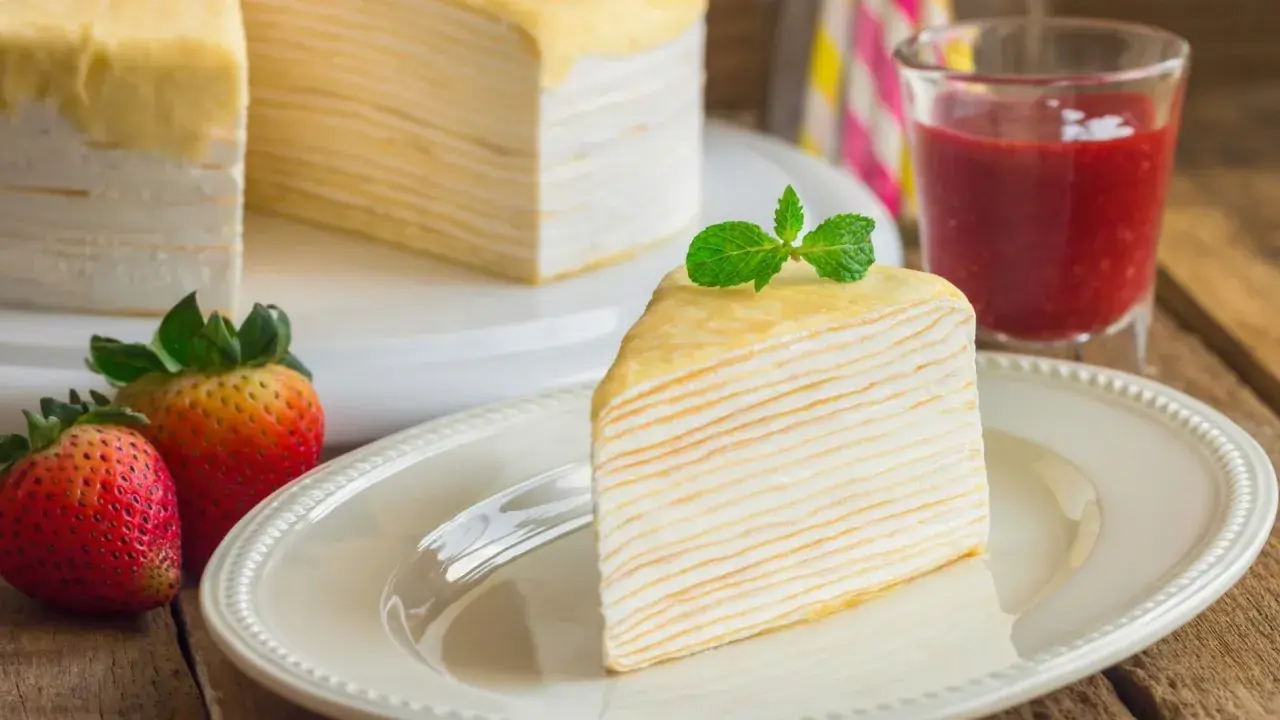 Una porción de tarta de crepes, decorada con menta, junto a fresas y un vaso de batido de fresa.