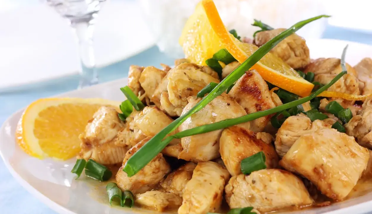 Sabroso pollo a la naranja, trozos jugosos con rodajas de naranja fresca y cebolleta picada, servido en un plato blanco.