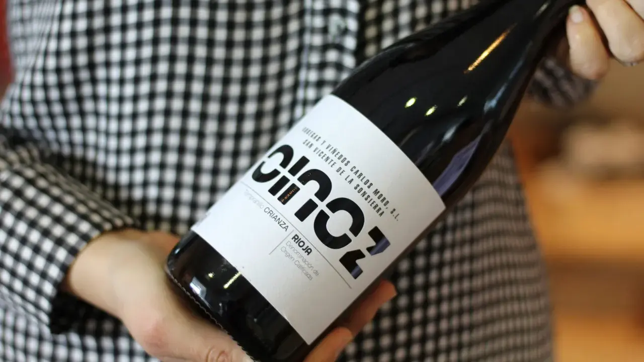 Manos sostienen una botella de vino blanco fermentado en barrica, con etiqueta negra y blanca, sobre un fondo de cuadros.