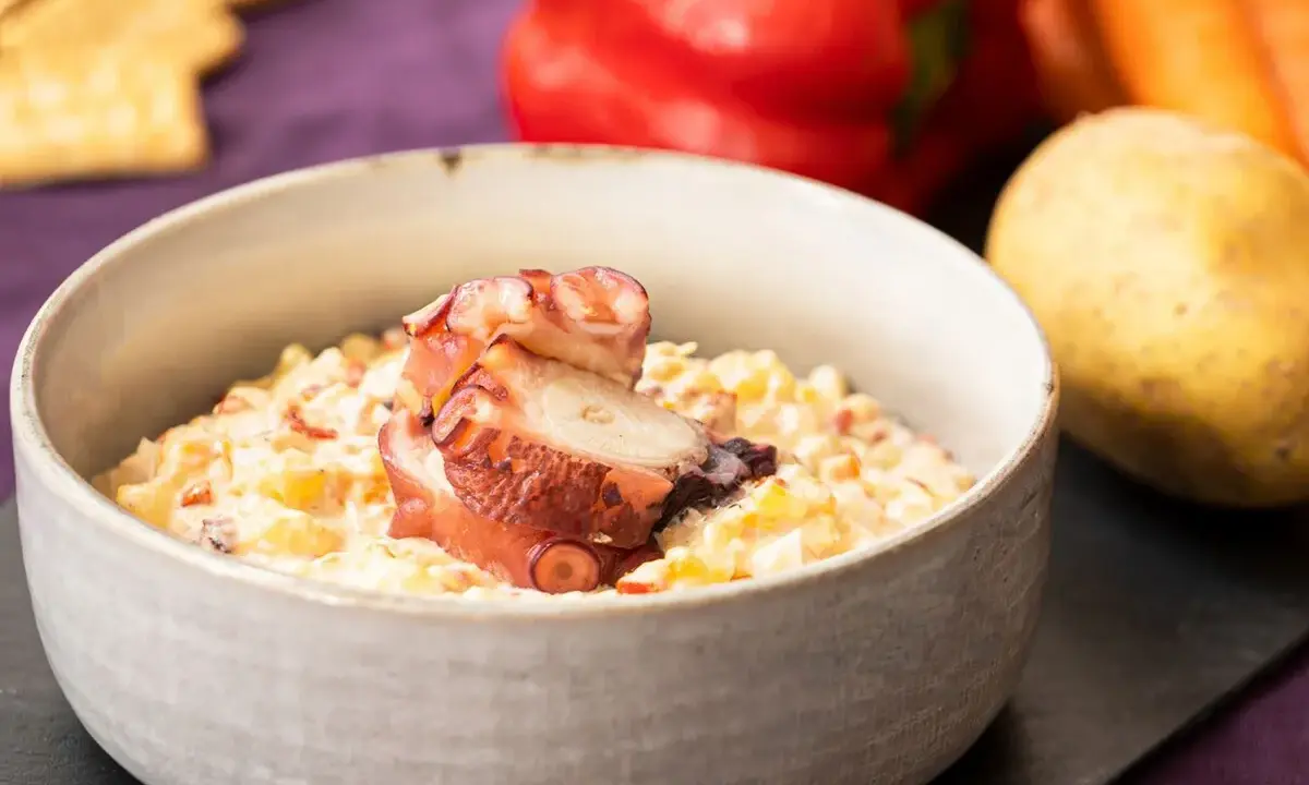 Deliciosa ensaladilla de pulpo cremosa, servida en cuenco, con patata, pimiento y zanahoria de fondo.