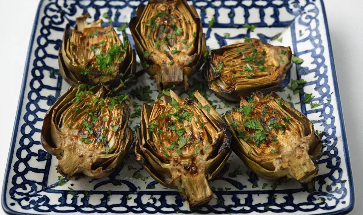 Seis mitades de alcachofas a la plancha, doradas y espolvoreadas con perejil fresco, servidas en un plato decorado.