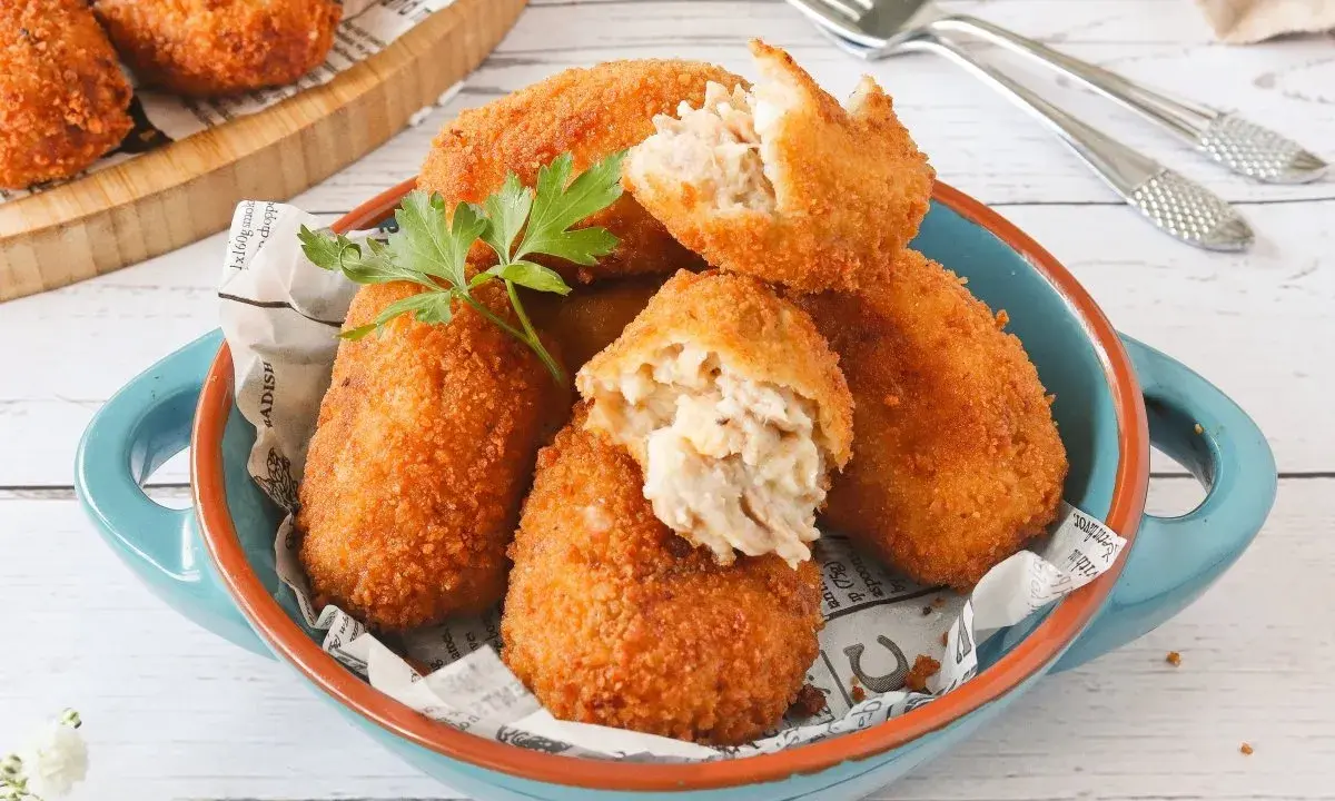 Croquetas de atún doradas y crujientes, servidas en un cuenco azul. Una está partida, mostrando su cremoso interior.