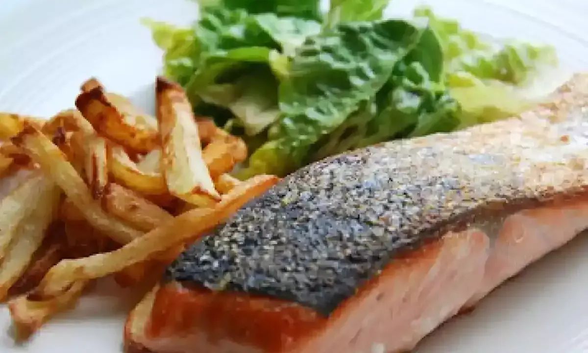Un jugoso salmón a la plancha con piel crujiente, acompañado de papas fritas doradas y una fresca ensalada verde.