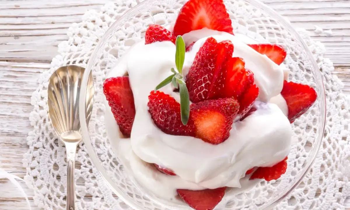 Delicioso postre de fresas con nata, servido en copa de cristal sobre un mantel de encaje. Una cucharilla plateada acompaña.