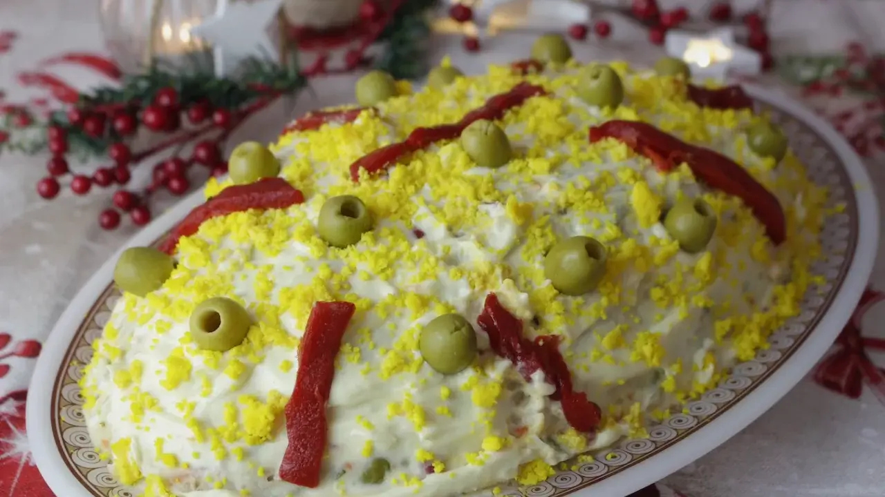 Ensaladilla rusa, la receta de la abuela, cubierta de mayonesa, huevo rallado, aceitunas y pimientos. ¡Un clásico festivo!