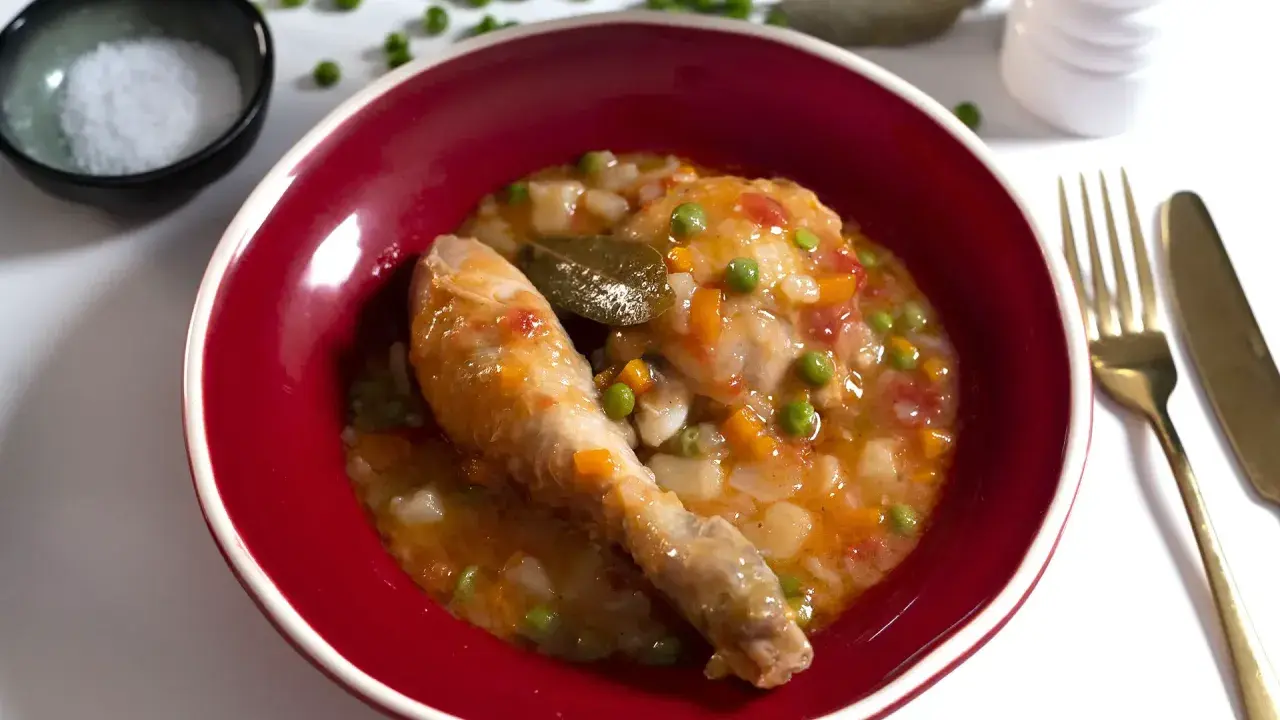 Un delicioso pollo a la jardinera en un cuenco rojo, con guisantes, zanahoria y tomate. Acompañado de sal, laurel y cubiertos dorados.