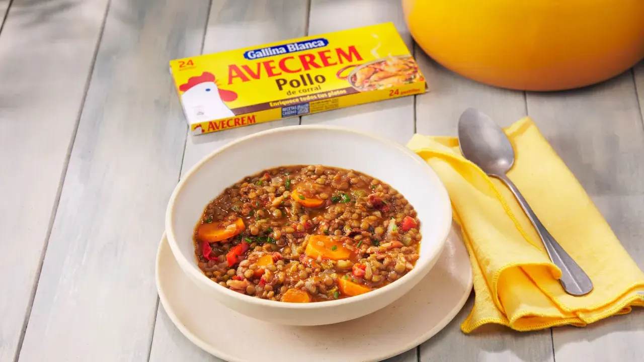 Un tazón de lentejas con zanahoria y pimiento rojo, listo para servir. La caja de Avecrem Pollo de corral, clave en esta lentejas receta de la abuela, descansa al lado.