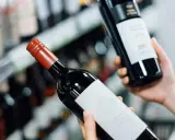Manos sostienen dos botellas de vinos caros, una con etiqueta blanca y otra con etiqueta negra, en un pasillo de supermercado.