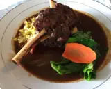 Delicioso lechazo guisado con hueso, acompañado de maíz tierno, zanahoria y verduras verdes en un plato blanco.