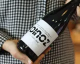 Manos sostienen una botella de vino blanco fermentado en barrica, con etiqueta negra y blanca, sobre un fondo de cuadros.