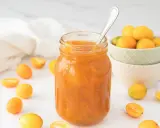 Tarro de mermelada de kumquat casera, rodeado de kumquats frescos y cortados. Una cuchara plateada reposa en el tarro.