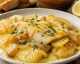 Merluza al horno con patatas, cebolla y perejil, servida en un plato blanco con una rodaja de limón al fondo.