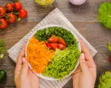 Manos sostienen un plato con una colorida ensalada mediterránea: zanahoria rallada, lechuga, perejil picado y tomates.
