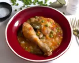 Un delicioso pollo a la jardinera en un cuenco rojo, con guisantes, zanahoria y tomate. Acompañado de sal, laurel y cubiertos dorados.