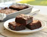Deliciosos trozos de brownie sin gluten apilados en un plato blanco, con más en un molde al fondo.