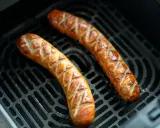 Dos salchichas doradas con cortes en forma de rombos, cocinándose en la rejilla de una freidora de aire.