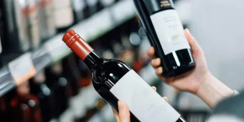 Manos sostienen dos botellas de vinos caros, una con etiqueta blanca y otra con etiqueta negra, en un pasillo de supermercado.