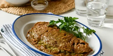 Tortilla de alcachofas dorada y jugosa, servida en plato blanco con borde azul. Decorada con perejil fresco.