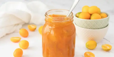 Tarro de mermelada de kumquat casera, rodeado de kumquats frescos y cortados. Una cuchara plateada reposa en el tarro.