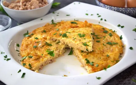 Tortilla de atún dorada y esponjosa, espolvoreada con perejil fresco. Un trozo cortado revela la textura jugosa.