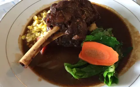 Delicioso lechazo guisado con hueso, acompañado de maíz tierno, zanahoria y verduras verdes en un plato blanco.