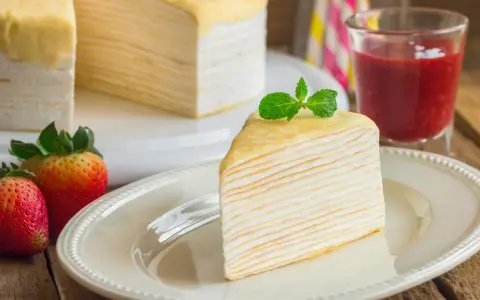 Una porción de tarta de crepes, decorada con menta, junto a fresas y un vaso de batido de fresa.