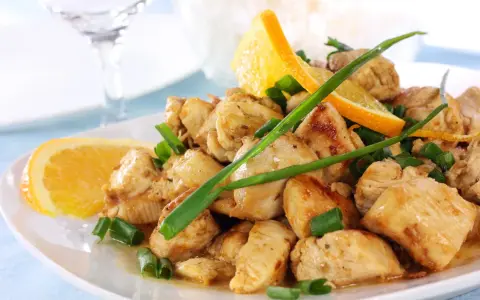 Sabroso pollo a la naranja, trozos jugosos con rodajas de naranja fresca y cebolleta picada, servido en un plato blanco.