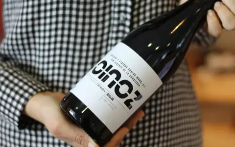 Manos sostienen una botella de vino blanco fermentado en barrica, con etiqueta negra y blanca, sobre un fondo de cuadros.