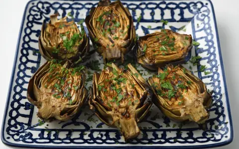 Seis mitades de alcachofas a la plancha, doradas y espolvoreadas con perejil fresco, servidas en un plato decorado.