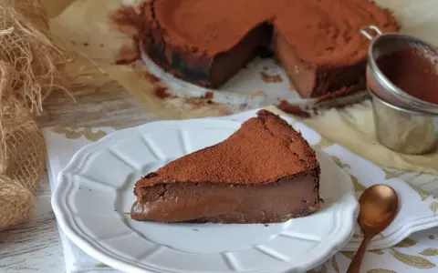 Un trozo de pastel de chocolate cubierto de cacao en polvo, con el pastel entero al fondo.