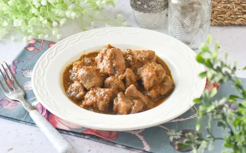 Plato de tierna carne de cerdo en salsa, servido en un cuenco blanco con un tenedor y decoración floral.