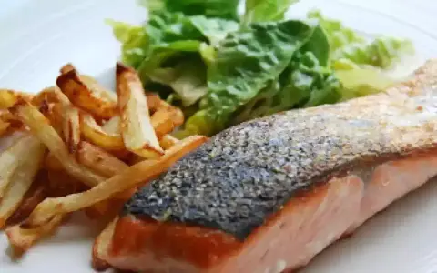 Un jugoso salmón a la plancha con piel crujiente, acompañado de papas fritas doradas y una fresca ensalada verde.