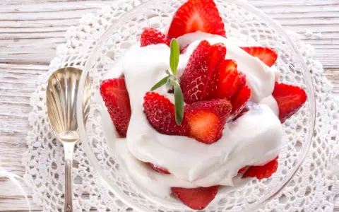 Delicioso postre de fresas con nata, servido en copa de cristal sobre un mantel de encaje. Una cucharilla plateada acompaña.