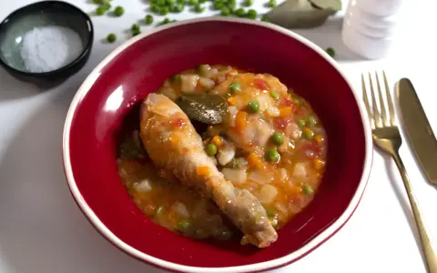 Un delicioso pollo a la jardinera en un cuenco rojo, con guisantes, zanahoria y tomate. Acompañado de sal, laurel y cubiertos dorados.