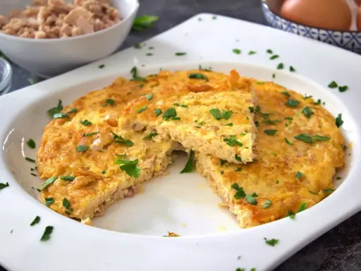 Tortilla de atún dorada y esponjosa, espolvoreada con perejil fresco. Un trozo cortado revela la textura jugosa.
