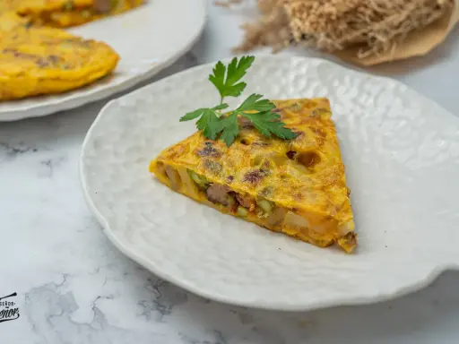 Un trozo de tortilla paisana, con patatas, verduras y chorizo, servida en un plato blanco texturizado.