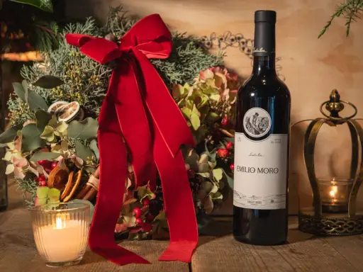 Botella de vino tinto fino Emilio Moro junto a un arreglo floral navideño, vela encendida y un candelabro dorado.