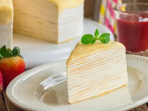 Una porción de tarta de crepes, decorada con menta, junto a fresas y un vaso de batido de fresa.