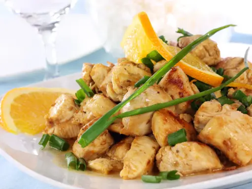 Sabroso pollo a la naranja, trozos jugosos con rodajas de naranja fresca y cebolleta picada, servido en un plato blanco.