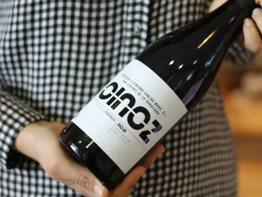 Manos sostienen una botella de vino blanco fermentado en barrica, con etiqueta negra y blanca, sobre un fondo de cuadros.