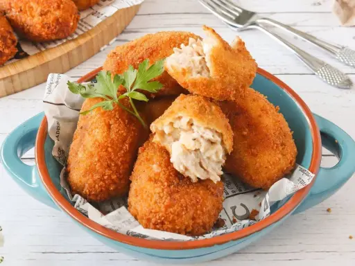 Croquetas de atún doradas y crujientes, servidas en un cuenco azul. Una está partida, mostrando su cremoso interior.