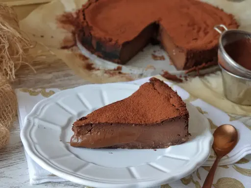 Un trozo de pastel de chocolate cubierto de cacao en polvo, con el pastel entero al fondo.