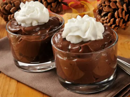 Dos copas de cremosas natillas de chocolate con nata montada, decoradas con piñas y hojas de otoño.