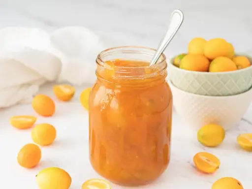 Tarro de mermelada de kumquat casera, rodeado de kumquats frescos y cortados. Una cuchara plateada reposa en el tarro.