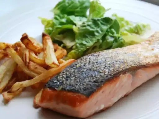Un jugoso salmón a la plancha con piel crujiente, acompañado de papas fritas doradas y una fresca ensalada verde.