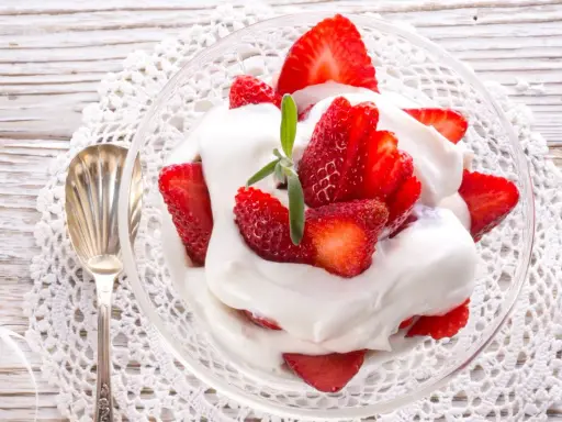 Delicioso postre de fresas con nata, servido en copa de cristal sobre un mantel de encaje. Una cucharilla plateada acompaña.