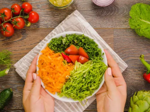 Manos sostienen un plato con una colorida ensalada mediterránea: zanahoria rallada, lechuga, perejil picado y tomates.