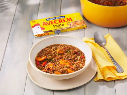 Un tazón de lentejas con zanahoria y pimiento rojo, listo para servir. La caja de Avecrem Pollo de corral, clave en esta lentejas receta de la abuela, descansa al lado.