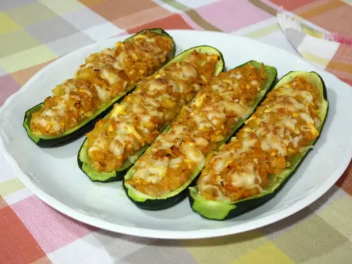 Cuatro calabacines rellenos de atún, gratinados con queso, servidos en un plato blanco sobre un mantel a cuadros.
